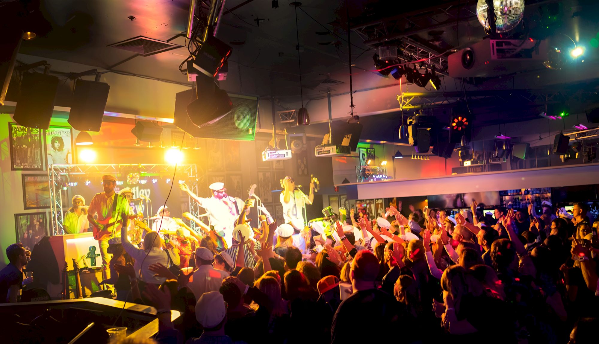 A live concert stage packed with musicians, colorful spotlights, and a cheering crowd raising hands.