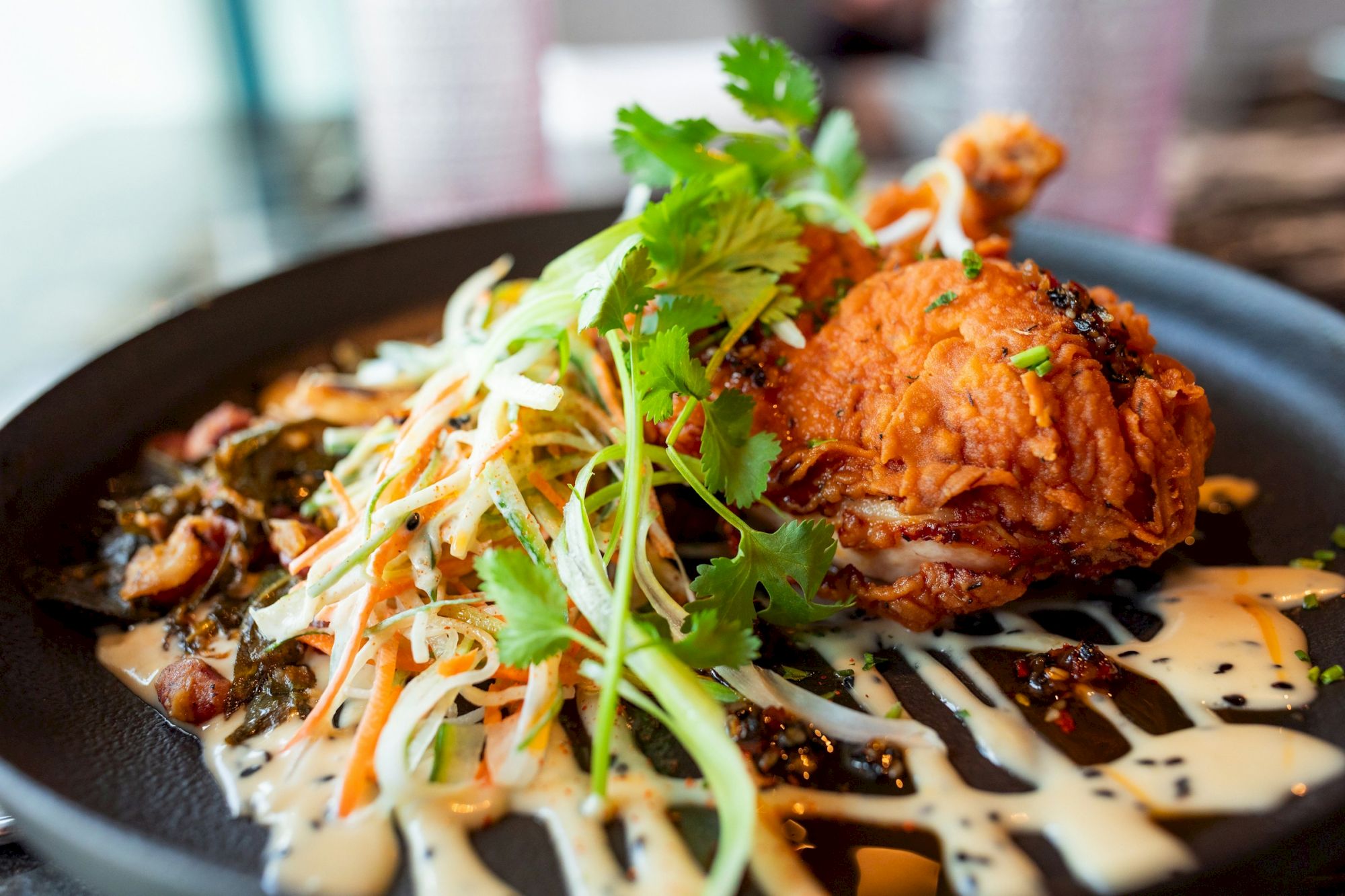 A plated fried chicken cutlet with shredded salad slaw, carrots, cilantro, and sauce on a dark plate.