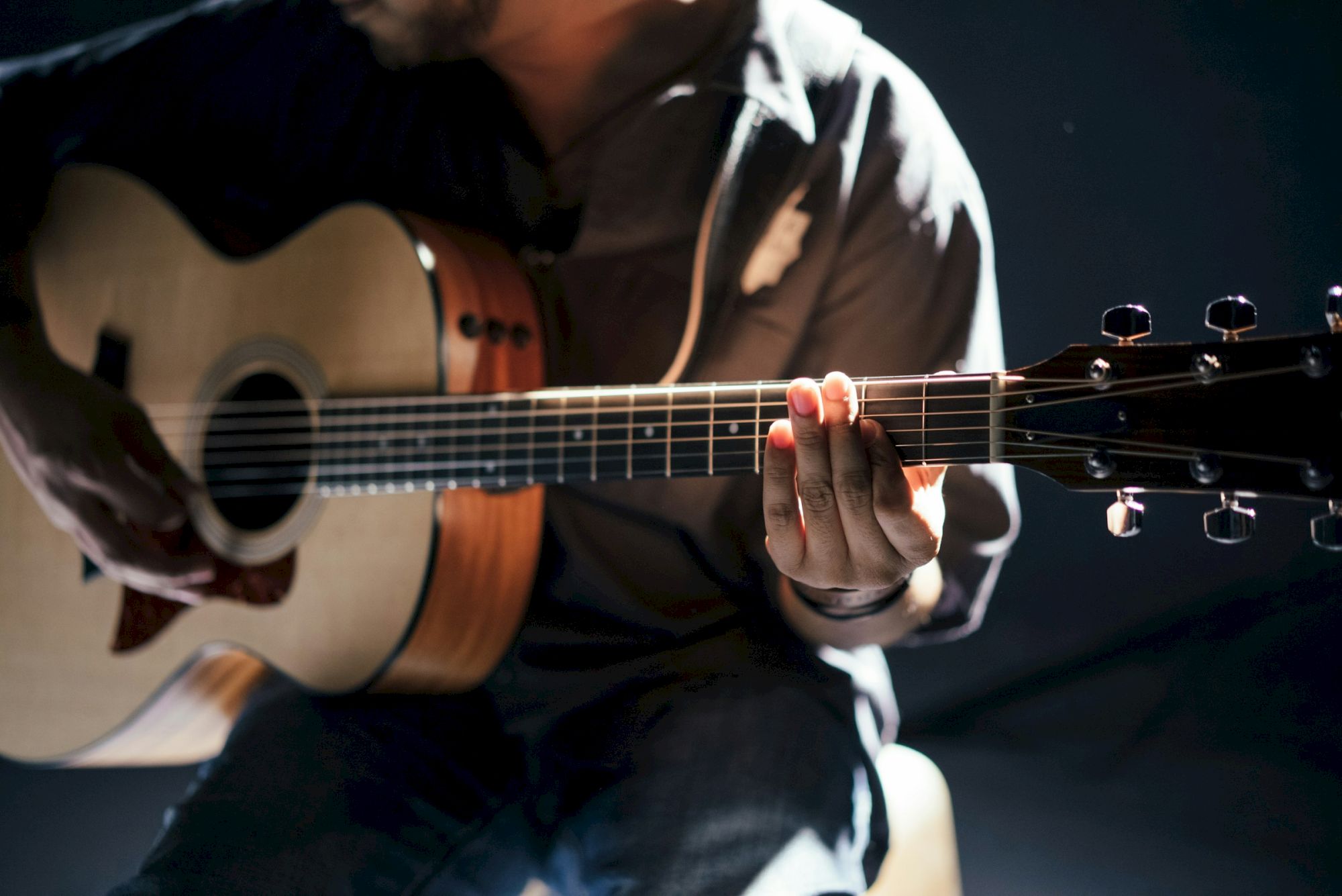A musician is playing an acoustic guitar in close-up, fingers pressing the strings under stage lights.
