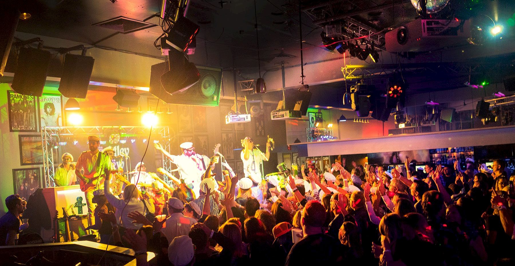 A live rock band performs on a brightly lit stage as a packed crowd cheers under colorful spotlights.