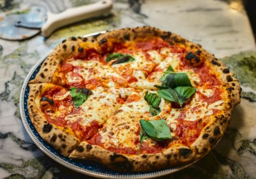 A wood-fired margherita-style pizza with melted cheese, red sauce, and fresh basil on a round pan.