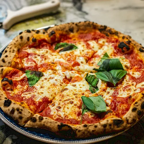 A wood-fired margherita-style pizza with melted cheese, red sauce, and fresh basil on a round pan.