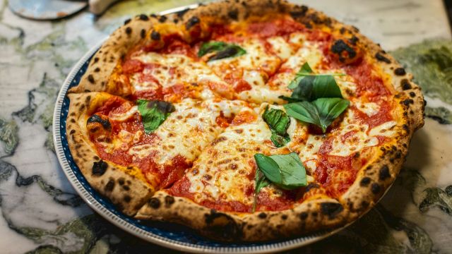 A wood-fired margherita-style pizza with melted cheese, red sauce, and fresh basil on a round pan.
