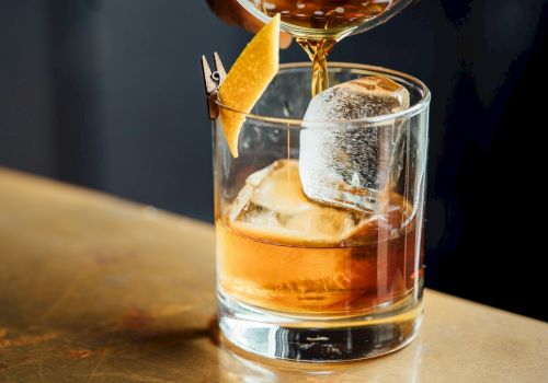 A bar pours amber liquor from a strainer into a glass over ice, with a lemon slice garnish floating upward.