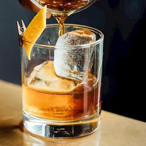 A bar pours amber liquor from a strainer into a glass over ice, with a lemon slice garnish floating upward.