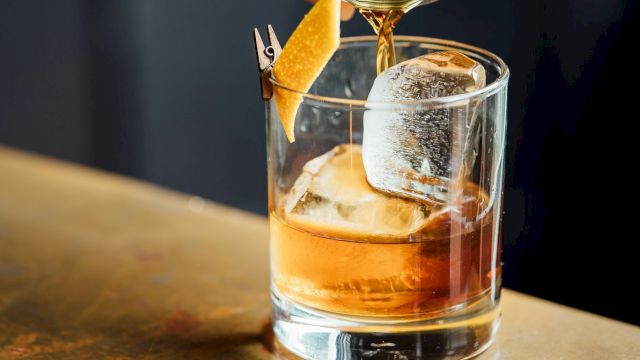 A bar pours amber liquor from a strainer into a glass over ice, with a lemon slice garnish floating upward.
