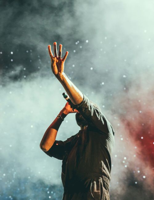A performer raises a hand in a smoky concert, with sparkling lights and drifting particles in the air.