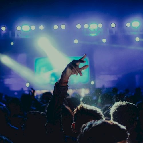A concert crowd cheers as a raised hand makes a peace sign under colorful stage lights.