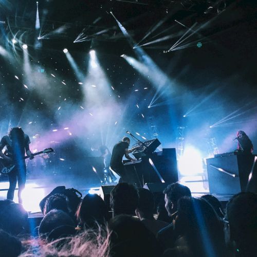 A live band performs on a dark stage under bright laser lights and fog, with a crowd watching in front.