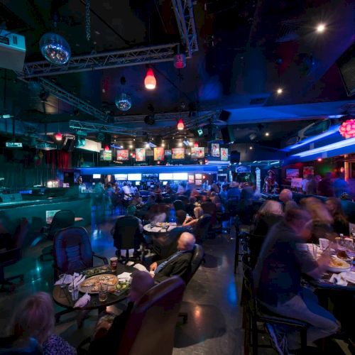 A lively nightclub/party hall with disco balls, a bar, and people dining under colorful lights.